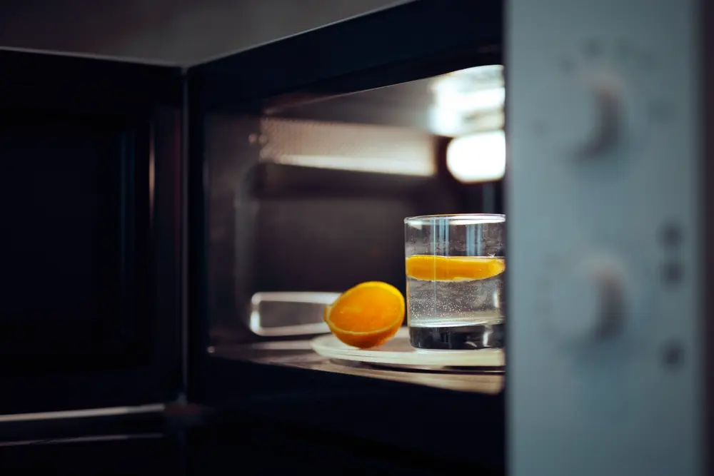 Copo com água e fatias de laranja dentro de um micro-ondas aberto, utilizado como método caseiro para eliminar odores.
