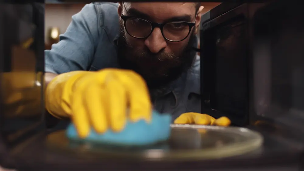 Homem usando luvas de borracha amarelas limpa o interior de um micro-ondas com uma esponja azul, visto de dentro do aparelho.