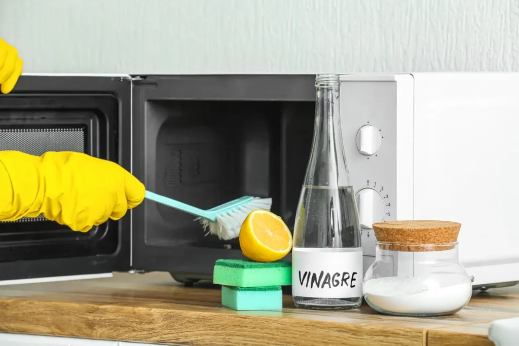 Pessoa com luvas de borracha amarelas limpando o interior de um micro-ondas com escova, enquanto sobre o balcão há meio limão, esponjas verdes, um pote de vidro com bicarbonato e uma garrafa de vidro transparente rotulada com a palavra 'Vinagre'.