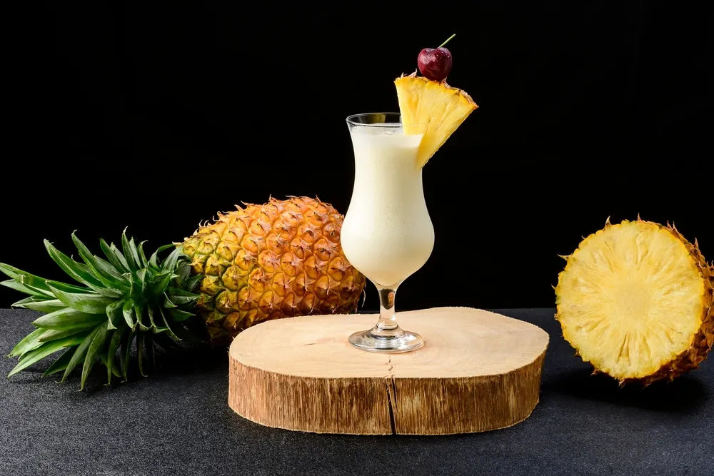 Copo de piña colada cremosa decorada com fatia de abacaxi e cereja, com abacaxi fresco ao fundo, bebida tropical clássica do Caribe.