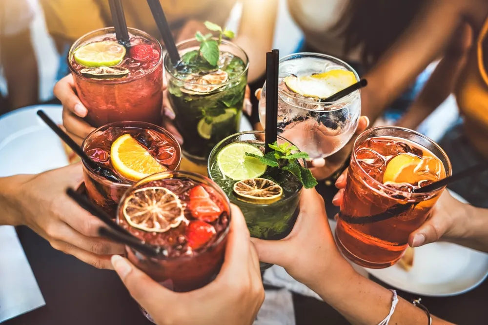 Grupo de amigos brindando com drinks coloridos sem álcool decorados com frutas cítricas, hortelã e gelo, em clima de celebração e verão.