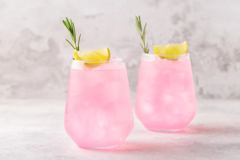 Dois copos de coquetel rosa com gelo, limão e ramo de alecrim, bebida refrescante e sofisticada servida em taça baixa.