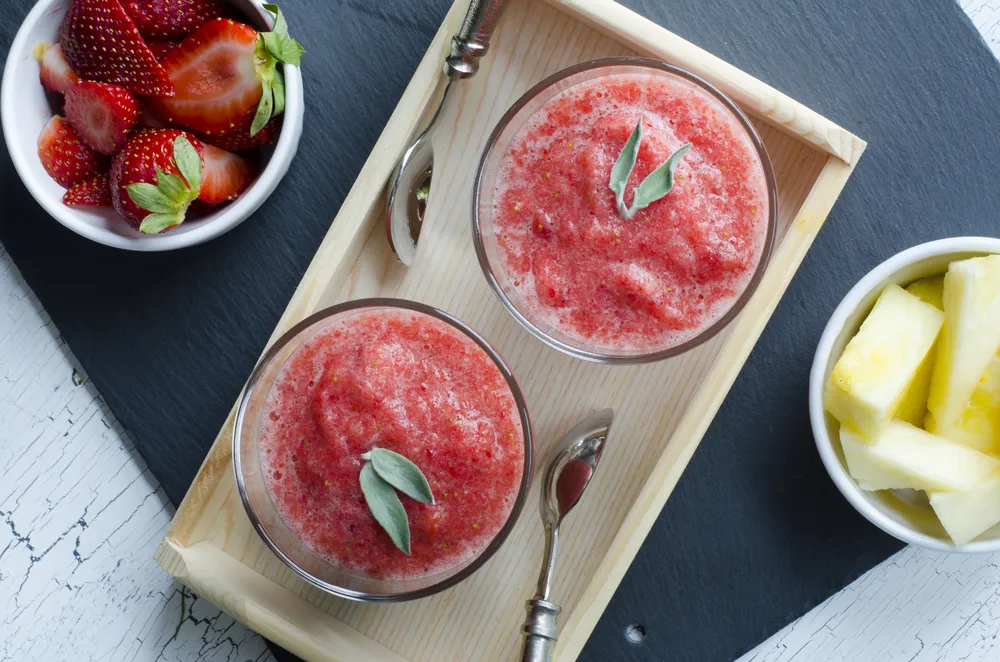 Batida tropical de morango servido em taças de vidro com folhas decorativas, acompanhada de frutas frescas como morangos e abacaxi.