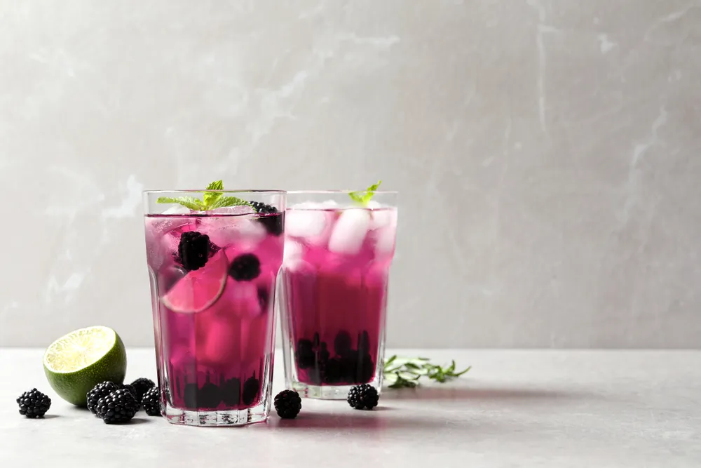 Bebida refrescante de amora e limão com gelo servida em copos transparentes, decorada com folhas de hortelã sobre fundo claro.