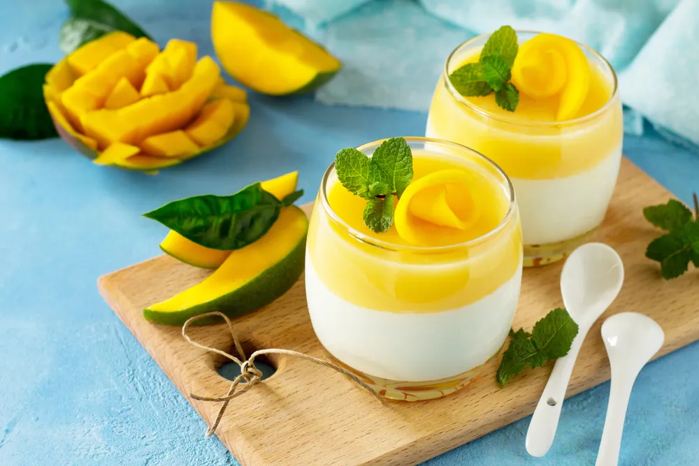 Smoothie de manga e coco servida em copos de vidro, decorada com hortelã e fatias de manga sobre tábua de madeira em fundo azul.