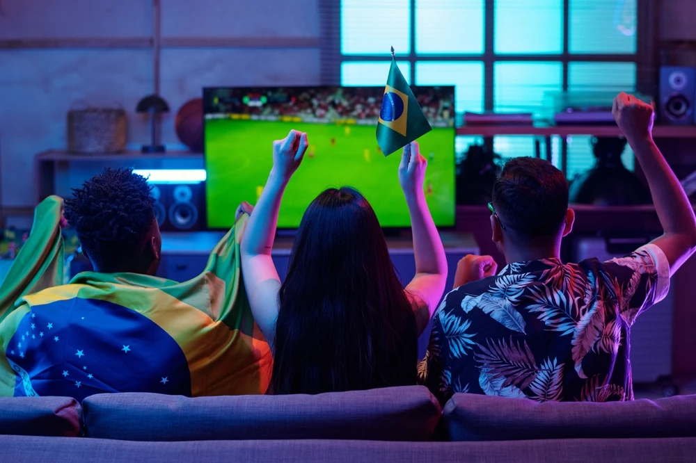 Grupo multiétnico de jovens adultos sentados em sofá assistindo a partida de futebol na televisão, levantando braços e agitando a bandeira brasileira, comemorando gol durante evento esportivo